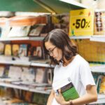 Woman shopping at an outdoor bookstore with a 50% discount sign, holding multiple books.